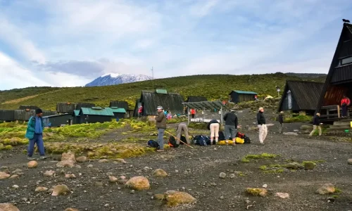 Hikers-are-preparing-gear-in-a-rocky-clearing-surrounded-by-dark-A-frame-huts-at-Horombo-Hut-with-the-snow-capped-peak-of-Kilimanjaro